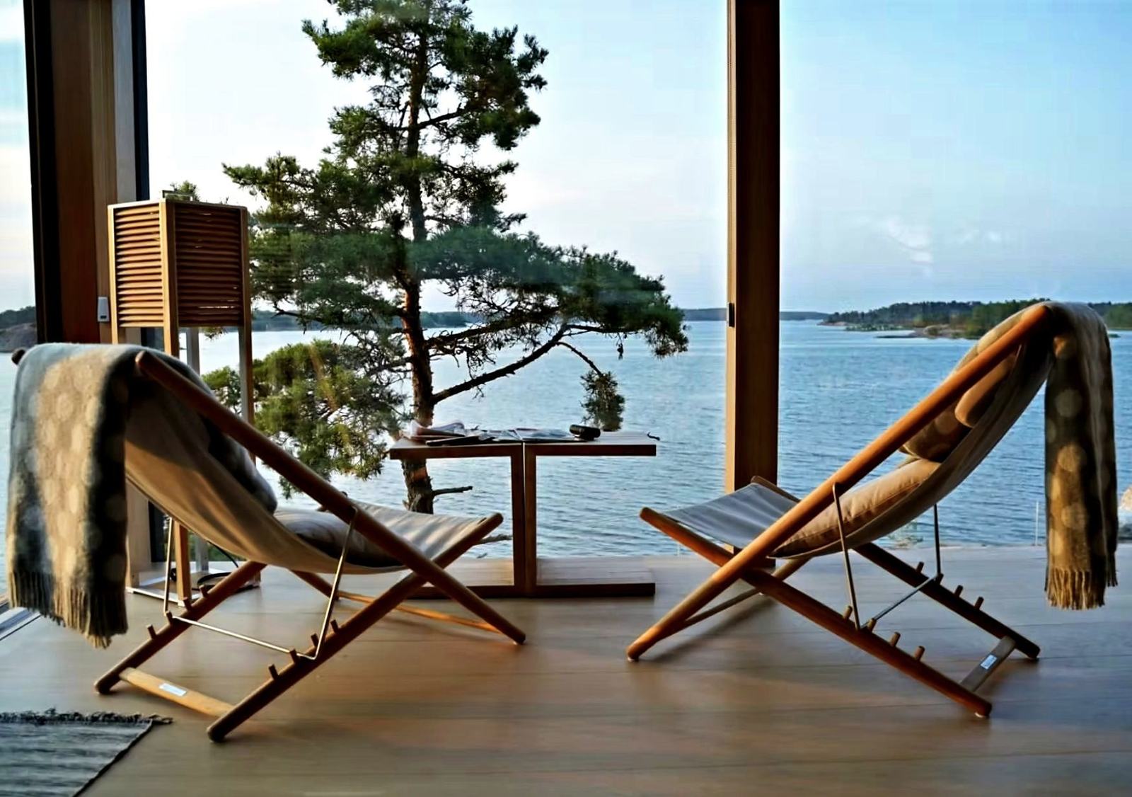 Two linen deck chairs at a wall of glass, a lone pine and the sea beyond
